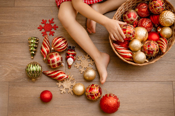 Dziecięce nóżki z koszykiem czerwonych i złotych zabawek świątecznych na drewnianej podłodze. Koncepcja świąteczna, przestrzeń tekstowa - Zdjęcie, obraz
