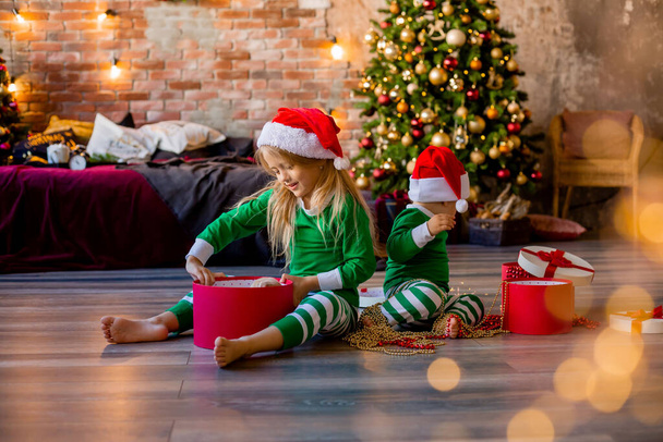 Zwei kleine Kinder in Pyjamas und Weihnachtsmützen packen im Zimmer Weihnachtsgeschenke aus - Foto, Bild