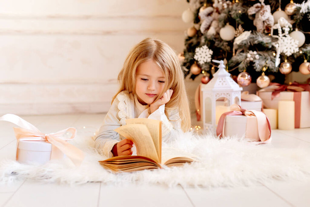 schattig klein blond meisje het lezen van een boek thuis in de buurt van de kerstboom - Foto, afbeelding