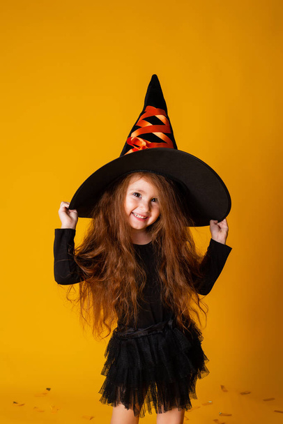klein meisje met lang rood haar in een Halloween heks kostuum op een gele achtergrond. Halloween voor kinderen, ruimte voor tekst - Foto, afbeelding