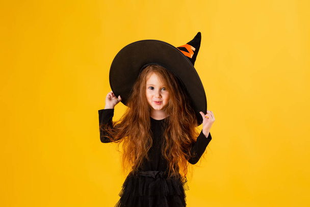 pieni tyttö, jolla on pitkät punaiset hiukset Halloween-noitapuvussa keltaisella taustalla. Halloween lapsille, tilaa tekstille - Valokuva, kuva