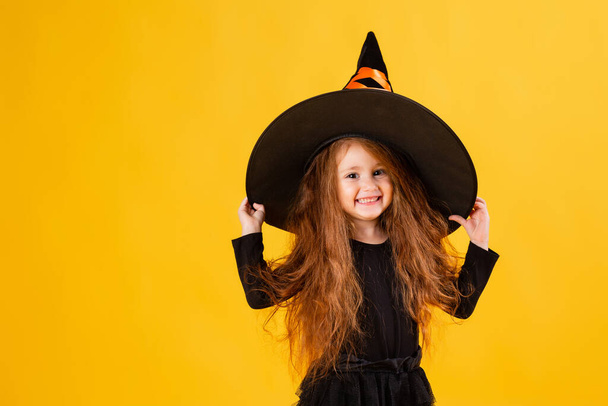 little girl with long red hair  in a Halloween witch costume on a yellow background. Halloween for children, space for text - 写真・画像