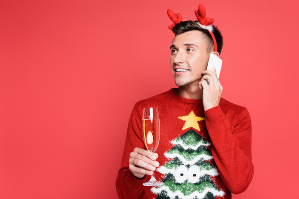 Homme souriant en pull avec pin parlant sur smartphone et tenant un verre de champagne isolé sur rouge - Photo, image