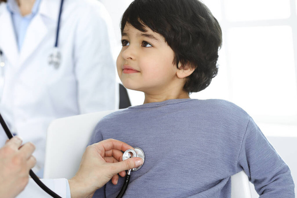 Doctor-woman examining a child patient by stethoscope. Cute arab boy at physician appointment. Medicine concept - Foto, afbeelding