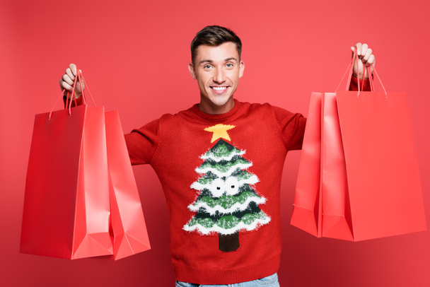 Sorrindo homem em suéter com pinheiro segurando sacos de compras e olhando para a câmera no fundo vermelho - Foto, Imagem
