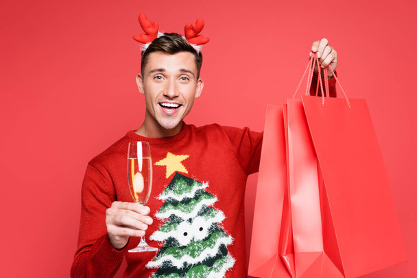 Mosolygó ember karácsonyi pulóverben kezében üveg pezsgő és bevásárló táskák elszigetelt piros - Fotó, kép