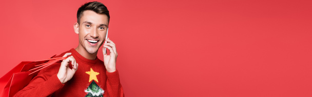 Young man in christmas sweater smiling while talking on smartphone and holding shopping bags isolated on red, banner - Foto, Imagem