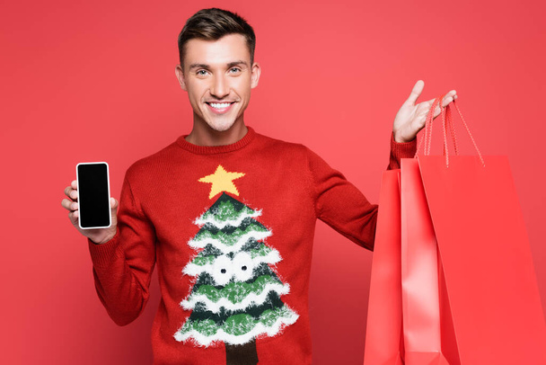 Homme souriant en pull avec pin tenant smartphone avec écran vierge et sacs à provisions sur fond rouge - Photo, image