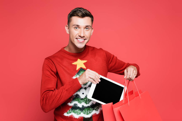 Uśmiechnięty mężczyzna w swetrze z choinką wkładający cyfrową tabletkę do torby na zakupy odizolowanej na czerwono - Zdjęcie, obraz