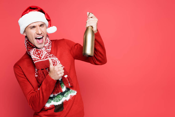 Excited man in santa hat showing like gesture and holding bottle of champagne on red background - Photo, image