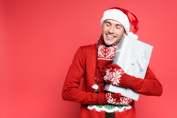 Smiling man in santa hat and mittens holding present on red background - Photo, Image