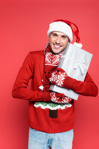 Cheerful man in santa hat and mittens looking at camera while holding gift box on red background - Photo, Image