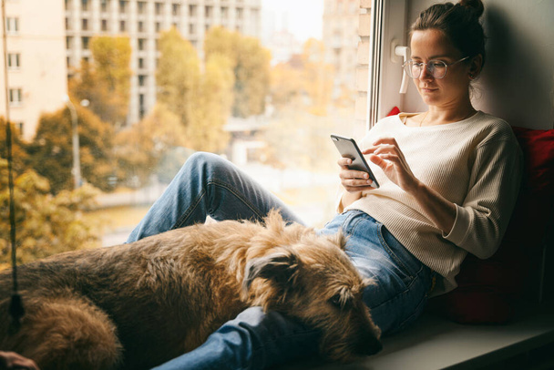 A young woman with her brown dog is sitting on a windowsill overlooking the city, looking at the smartphone screen stroking her pet - Foto, Bild