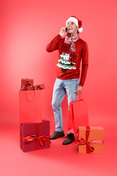Smiling man in santa hat talking on smartphone near shopping bags and gift boxes on red background - Foto, Imagem