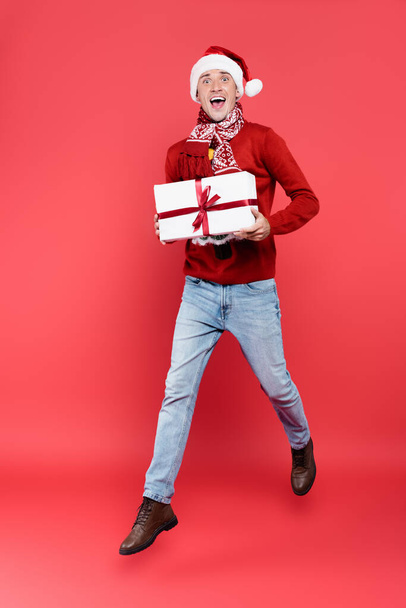 Cheerful man in santa hat and scarf holding present while jumping on red background - Photo, Image