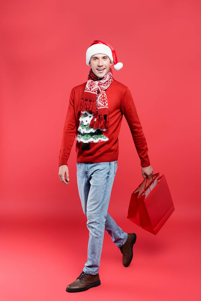 Smiling man in santa hat holding shopping bags while walking on red background - Φωτογραφία, εικόνα