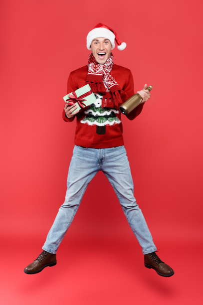 Excited man in scarf and santa hat holding gift box and bottle of champagne while jumping on red background - Foto, Imagen