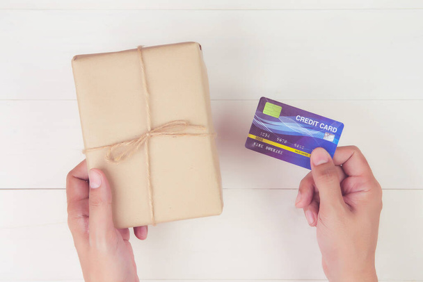 Hand eines Mannes mit Kreditkarte und Online-Einkauf zum Schenken von Geschenkschachteln mit Kraftpapier auf Holzgrund, Geschenk zur Feier auf dem Schreibtisch zu Weihnachten oder im Urlaub, von oben gesehen, flache Lage. - Foto, Bild