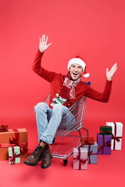 Hombre alegre en suéter y sombrero de santa sentado en el carrito de la compra cerca de regalos sobre fondo rojo - Foto, imagen
