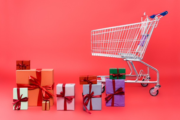 Gift boxes with ribbons near shopping cart on red background - Foto, Imagen