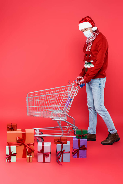 Man in santa hat and medical mask looking at gifts near shopping cart on red background - Foto, imagen