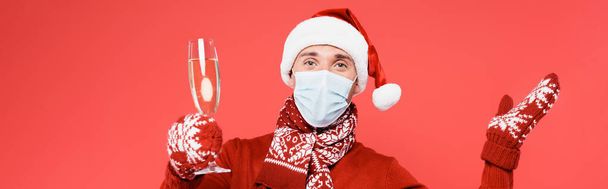 Uomo in cappello di Babbo Natale e maschera medica contenente bicchiere di champagne isolato su rosso, banner - Foto, immagini