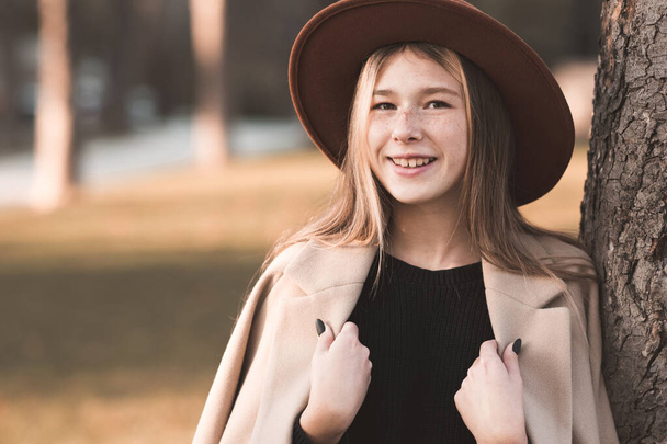 Посмішка дівчинки-підлітка 13-14 років, одягнена в стильний капелюх і бежеве зимове пальто, висить над осіннім фоном. Підлітковий період.  - Фото, зображення