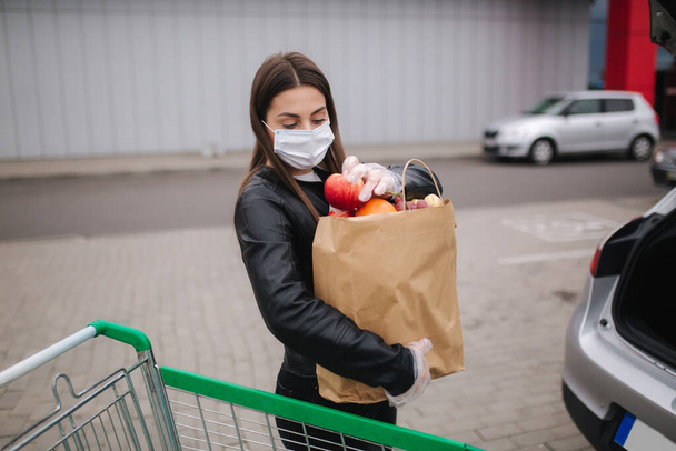 Koruyucu maskeli ve tek kullanımlık eldivenli genç bir kadın süpermarketin önündeki arabasına bir el işi paketi taşıyor. - Fotoğraf, Görsel