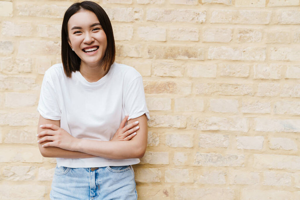 Picture of a happy young asian woman posing over wall outdoors - Foto, Bild