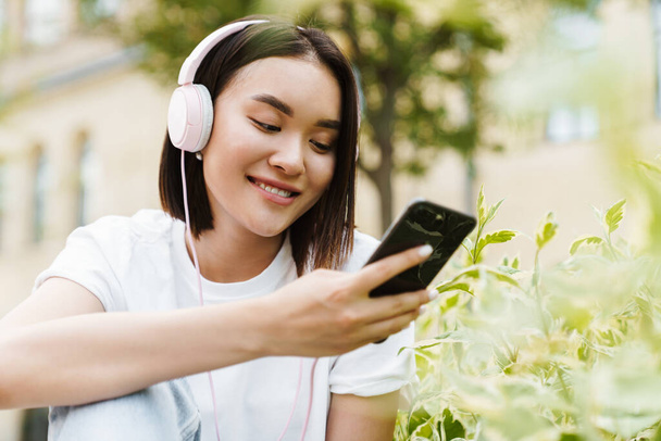 Изображение позитивной молодой азиатки, сидящей на улице, используя мобильный телефон и слушая музыку в наушниках - Фото, изображение