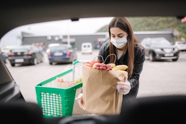 Over trunk view of young woman in medical mask in masks loading bags in trunk after supermarket shopping. Quqrantine grocery shipping. Fresh fruits for grandparents. Female with eco craft paket - Photo, Image