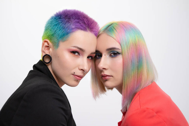 Young lesbian woman couple with vivd colored short hair and jackets posing on white background. Piercing on the face, tunnels in the ears. - Фото, зображення