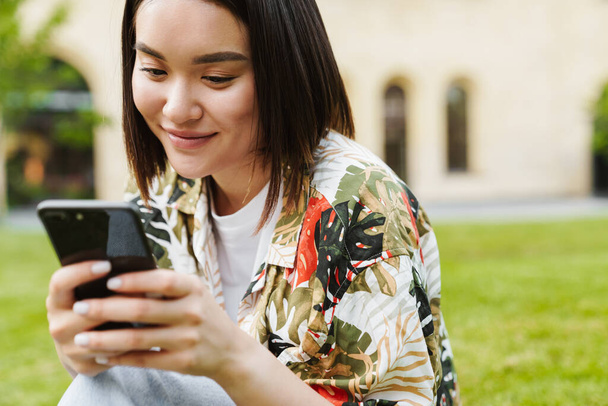 Photo of pleased young asian woman sitting outdoors on grass using mobile phone - Φωτογραφία, εικόνα