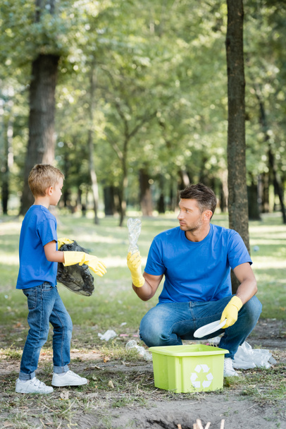 father and son collecting plastic garbage into container with recycling emblem, ecology concept - Fotografie, Obrázek