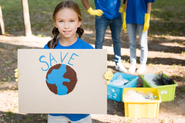 niña sosteniendo pancarta con globo y guardar la inscripción, y los padres cerca de contenedores de basura sobre fondo borroso, concepto de ecología - Foto, Imagen