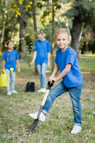 boy digging ground with shovel near mother and sister with watering can and young seedling on blurred background, ecology concept - Photo, Image