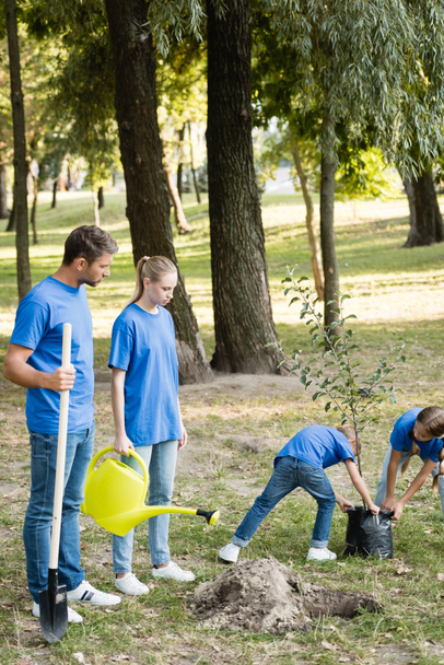 fiatal fát hordozó gyerekek, miközben a szülők ásóval és öntözőkannával állnak, ökológia koncepció - Fotó, kép