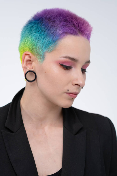 A young beautiful girl with short colored hair. Spread bright coloring and creative make-up. Piercing on the face. A black jacket. - Фото, изображение