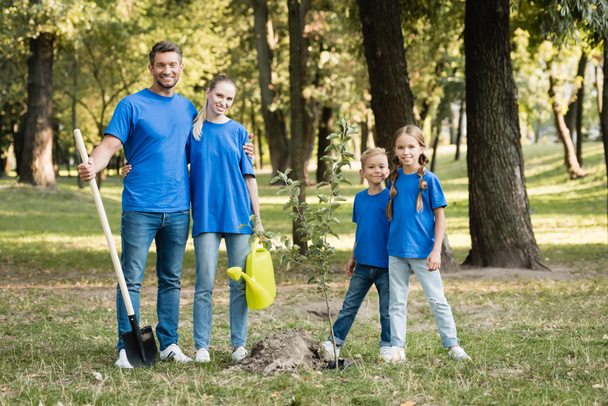 happy family of volunteers standing near planted young tree with shovel and watering can, ecology concept - Photo, Image