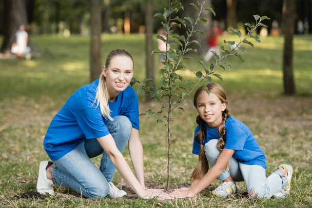 sonrientes madre e hija mirando a la cámara mientras plantan un árbol joven en el parque, concepto de ecología - Foto, imagen