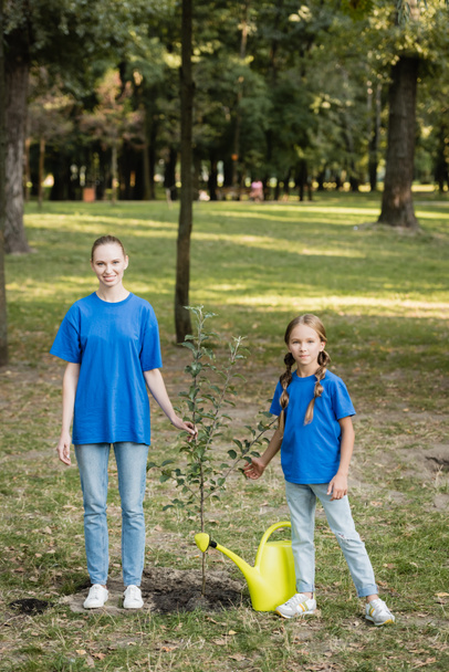 happy mother and daughter looking at camera while standing near young planted tree, ecology concept - Foto, imagen