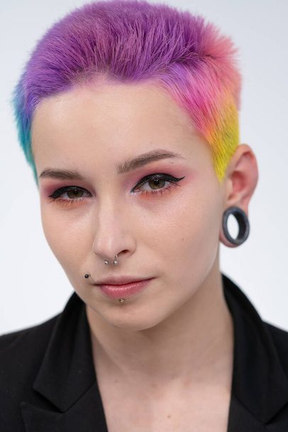 A young beautiful girl with short colored hair. Spread bright coloring and creative make-up. Piercing on the face. A black jacket. - Photo, Image