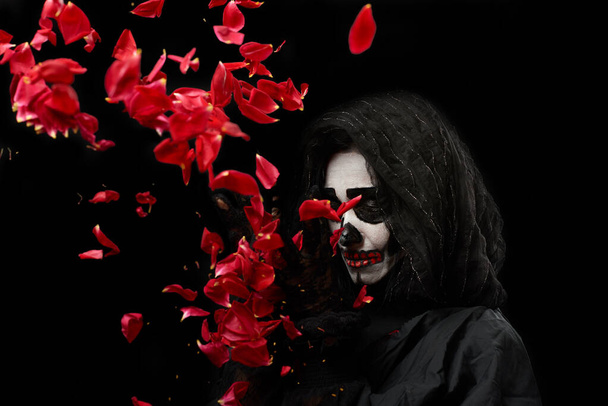kobieta z makijażu szkielet stoi w czarne ubrania i przezroczysty kaptur i wymiotuje czerwone róże na czarnym tle - Zdjęcie, obraz