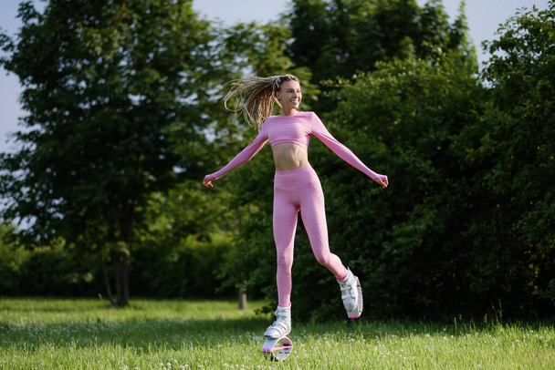 A young slender beautiful woman with dreadlocks in sportswear trains in the park outdoors. Kangoo jumps training. - Φωτογραφία, εικόνα
