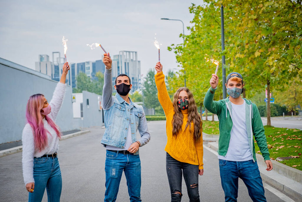 Group of multiethnic friends jocking with sparklers and wearing face safety masks while keeping social distance during coronavirus time - Covid-19 spread prevention concept - Foto, imagen