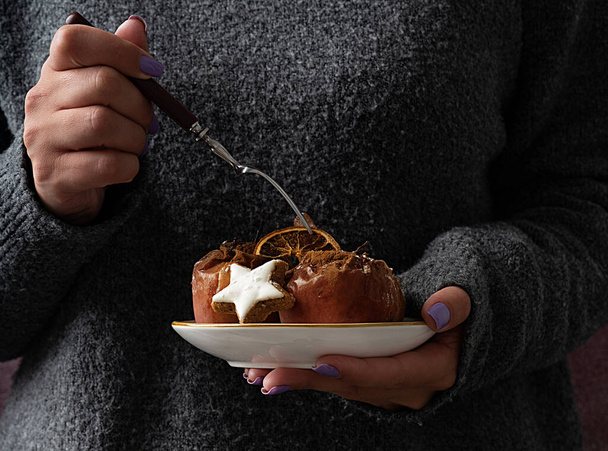 Female in grey knitted sweater holding a fork and ceramic plate with baked apples decorated with dry orange, Christmas star cookie and cinnamon powder. Christmas concept. Rustic style. - Photo, Image