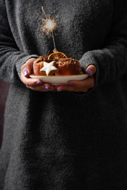 Mujer en suéter de punto gris sosteniendo un plato de cerámica con manzanas horneadas decoradas con naranja seca, galleta estrella christmast, espumoso y canela en polvo. Concepto de Navidad. Estilo rústico. - Foto, Imagen