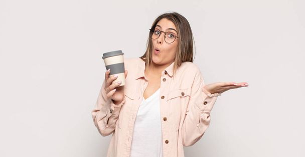 young blonde woman looking surprised and shocked, with jaw dropped holding an object with an open hand on the side - 写真・画像