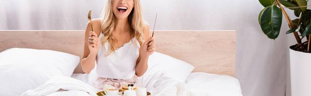 Cropped view of cheerful woman holding cutlery near breakfast on tray on bed, banner  - Photo, image