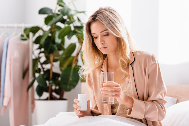 Young woman looking at jar with pills and holding glass of water on bed  - Foto, Bild